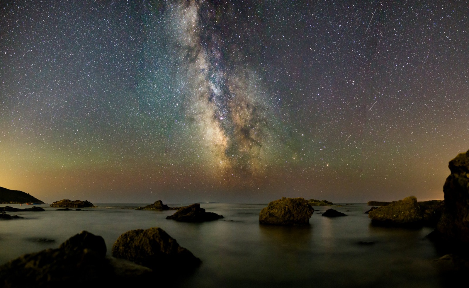 Milky Way rising over dark shoreline rocks and calm sea at night