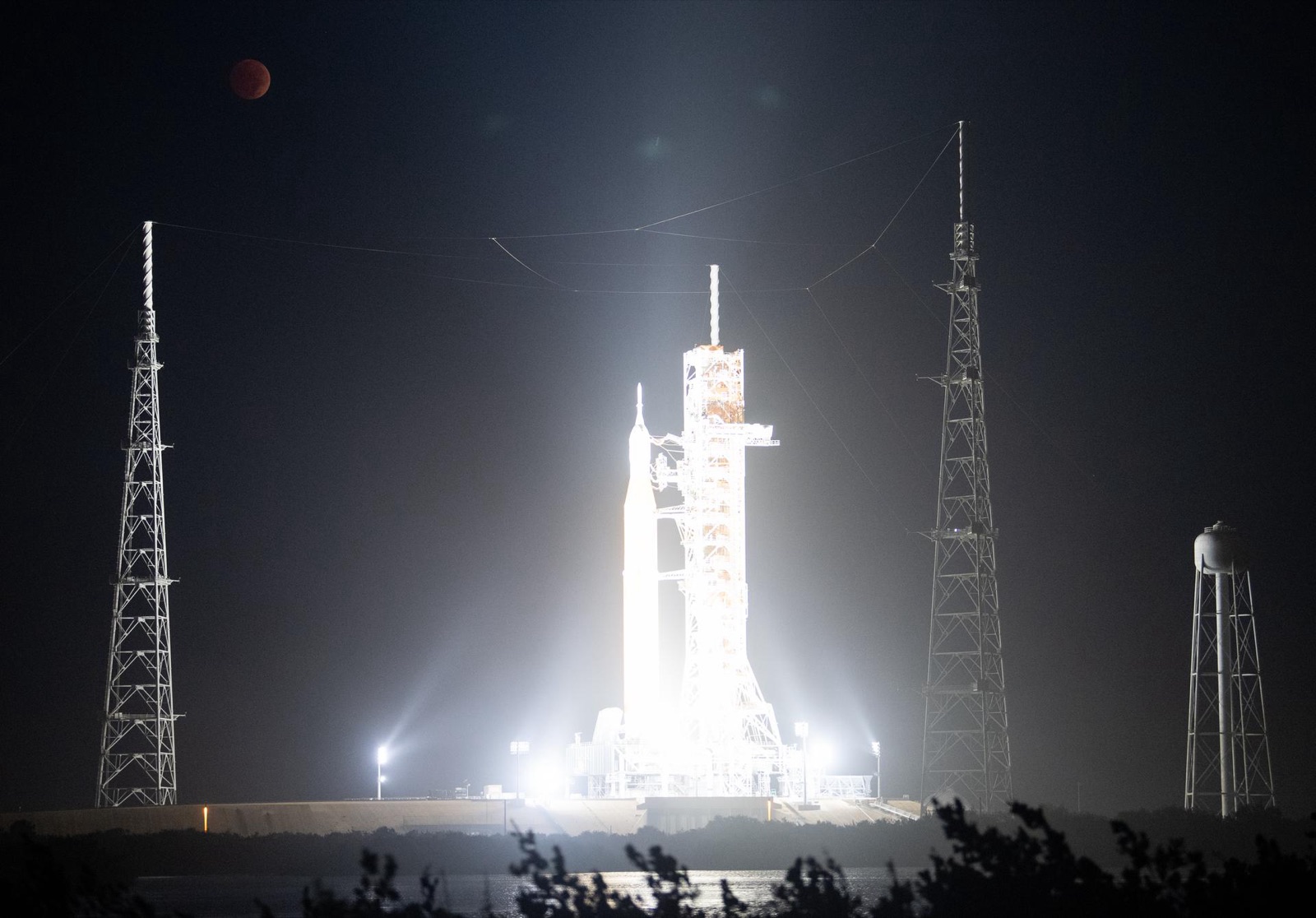 Small red eclipsed Moon at upper left above a floodlit rocket and launch towers at night.
