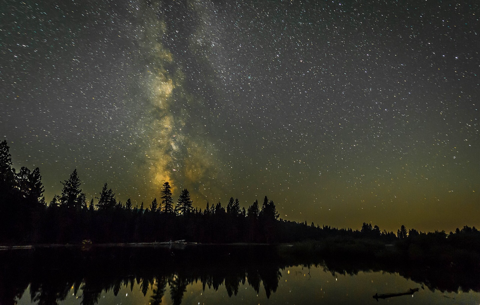 Milky Way core rising above silhouetted conifers with stars reflected in a still lake.