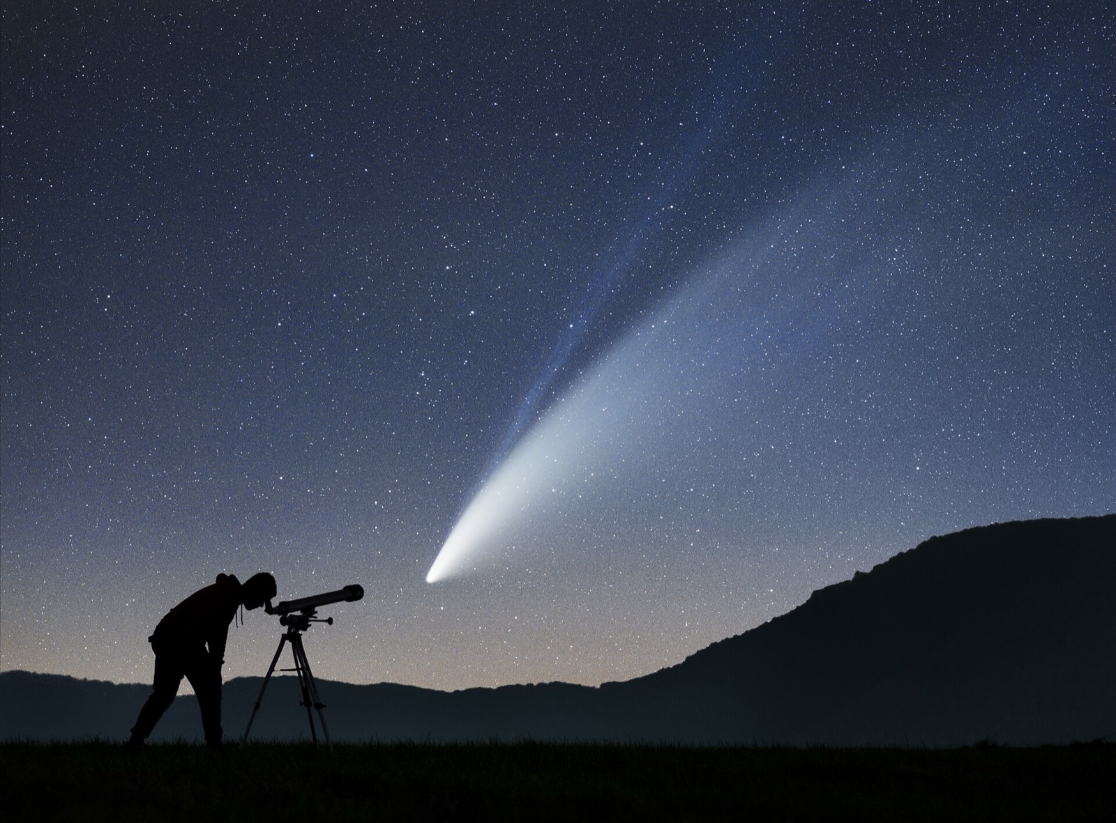Comet with a wide pale dust tail and thin blue ion tail above a hill and a silhouetted observer.