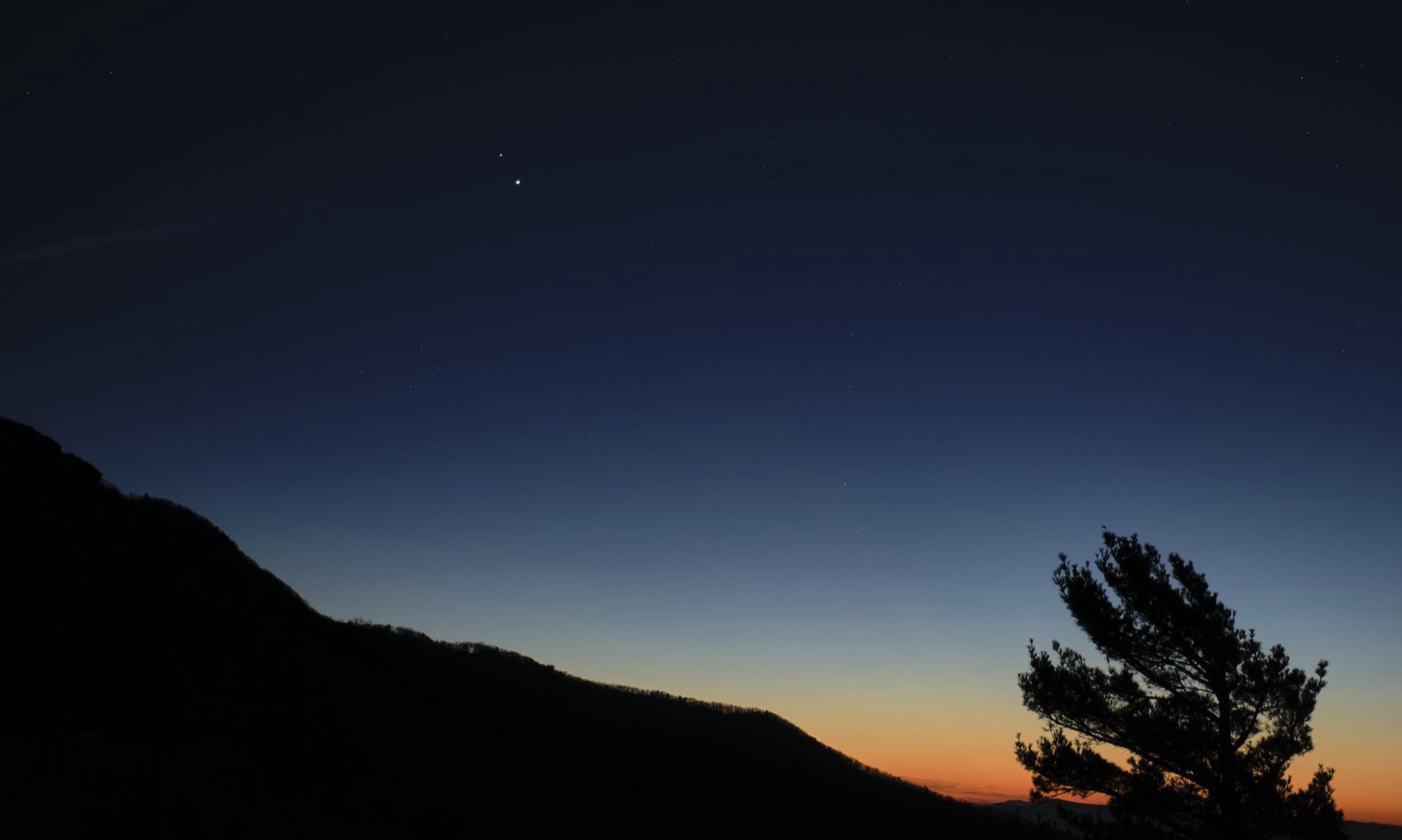 Two planets close together in a deep blue dusk sky above a dark ridge and a tree.