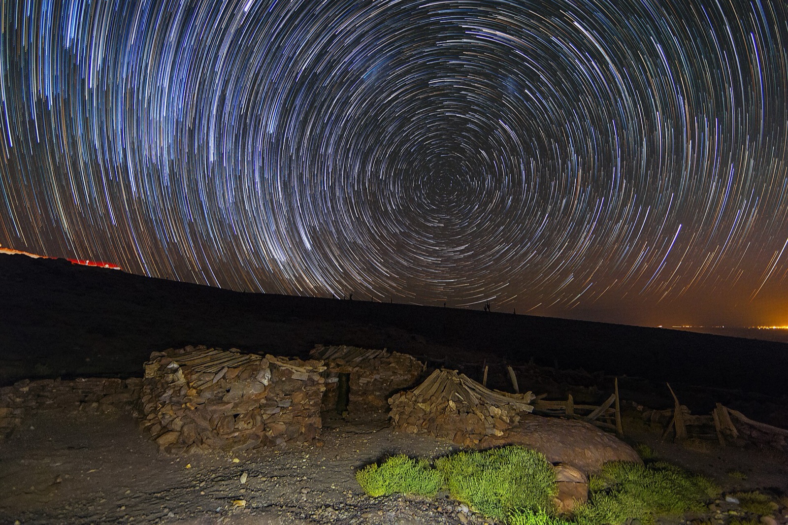 Curved star trails above a dry-stone wall and a hillside at night.