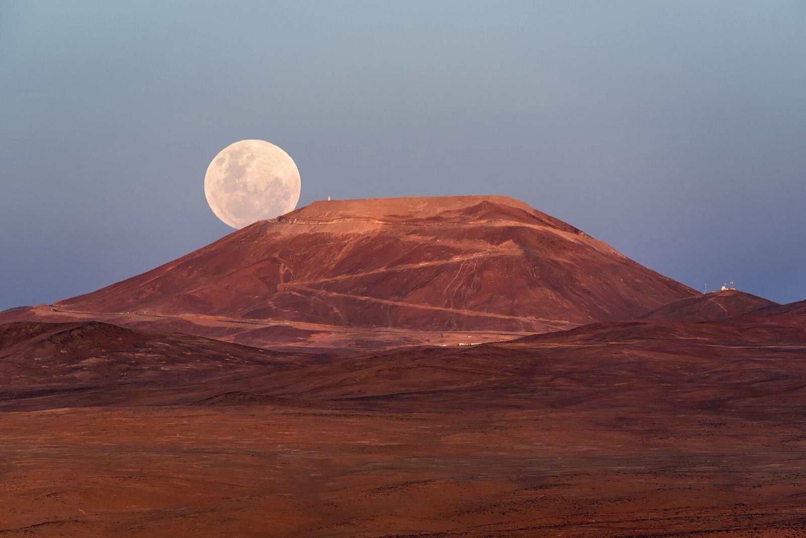 Full Moon rising behind a flat-topped desert mountain lit red by dusk under a pale sky.