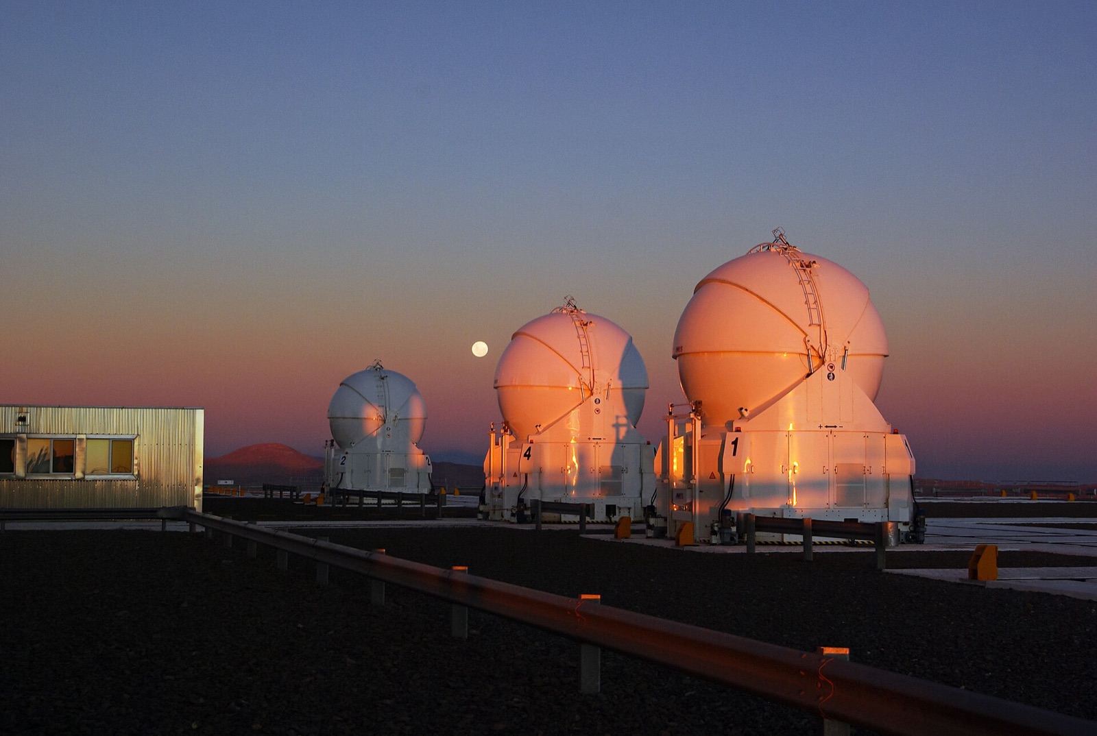 Pink band of sky above a blue-grey horizon band, with the Moon and orange-lit telescope domes.