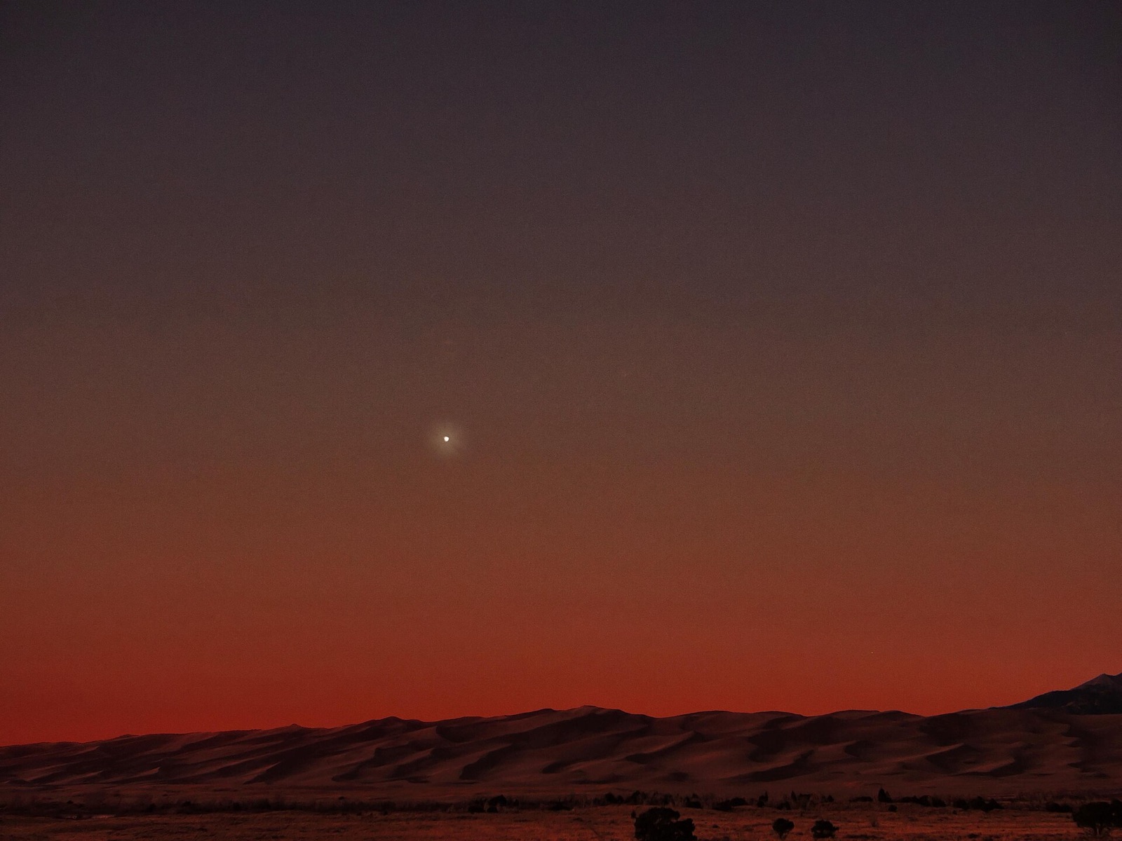 Bright Venus in a dark sky above a red twilight band and silhouetted dunes.